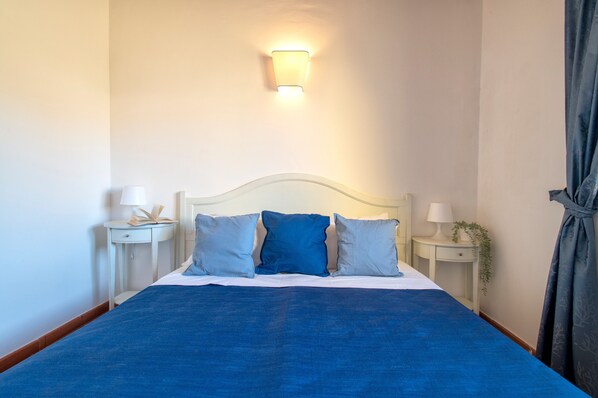 Un letto rifatto con una coperta e cuscini blu, due comodini con lampade, e un lampadario a parete.