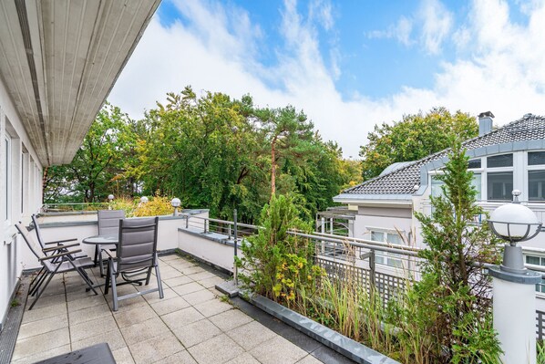 A rooftop terrace with outdoor seating, a railing, and a view of surrounding buildings and trees.
