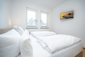 A bedroom with two windows, a bed with white bedding, and a wall-mounted picture of a sunset over water.