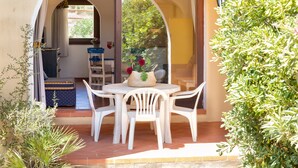 Een terras buiten met een ronde tafel en witte stoelen, een klein tapijt en een plant in pot.