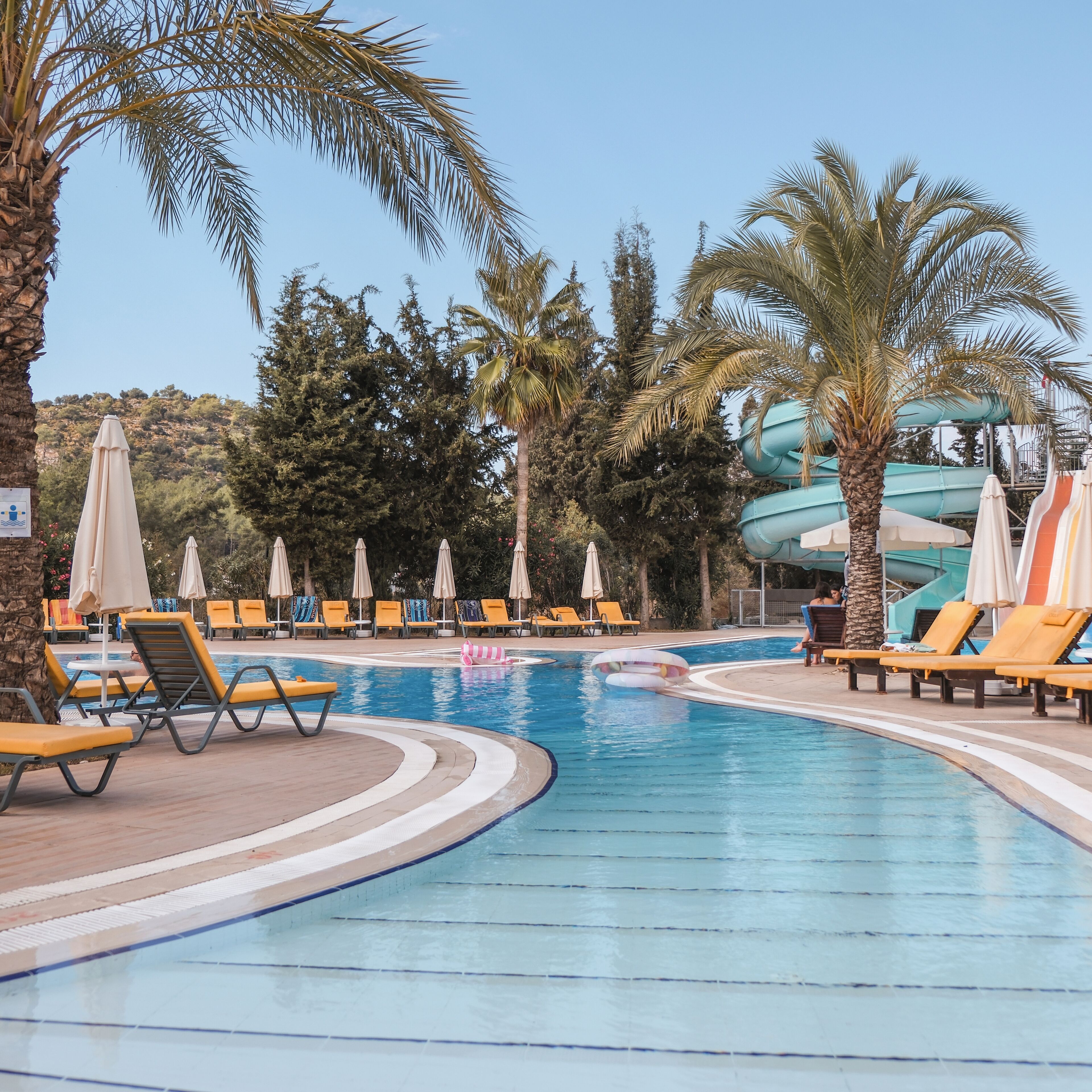 A pool area with lounge chairs and umbrellas, surrounded by palm trees and a water slide.