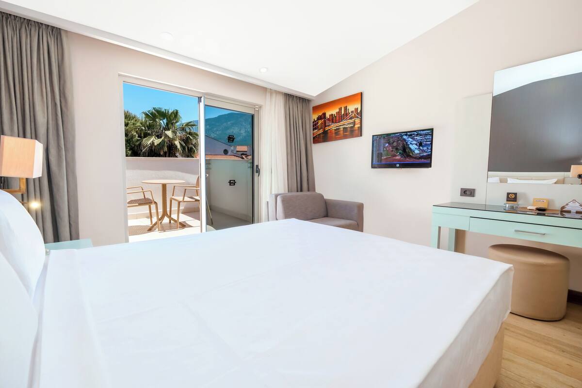 A hotel room with a bed, a desk, a chair, a TV, and a balcony with a view of palm trees and mountains.