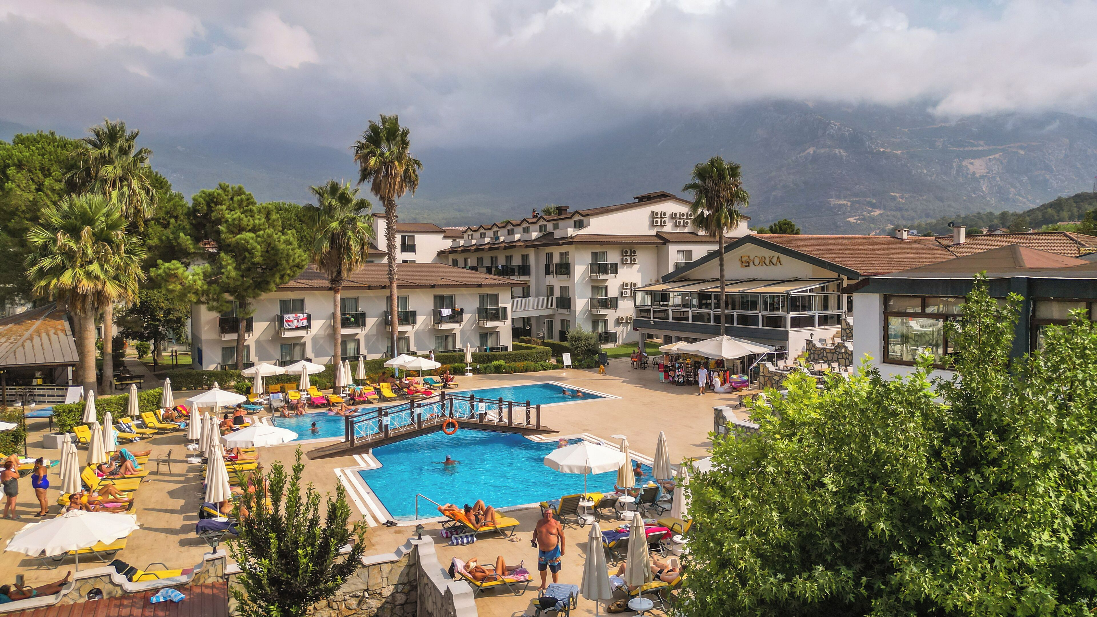 A resort with a swimming pool surrounded by sun loungers and umbrellas, a restaurant, and a view of mountains.