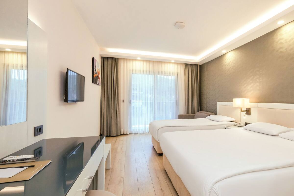 A hotel room with a large bed, a desk with a computer, a TV, and a window with curtains.