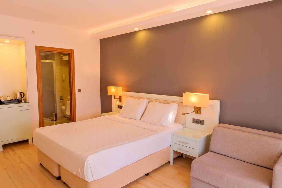 A modern hotel room with a large bed, bedside lamps, a sofa, and a wooden floor.