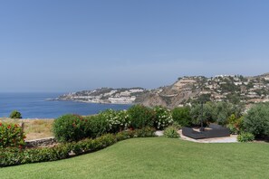 A coastal landscape with a grassy area, a patio with seating, and a view of the sea and distant buildings. - Villa Las Dalias Almunecar and Nerja (Almunecar)