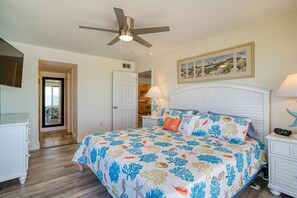 A bedroom with a bed, a ceiling fan, a painting, and a nightstand with a lamp.