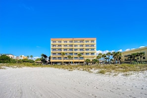 A multi-story hotel with balconies, located on a beach with palm trees and sandy ground.