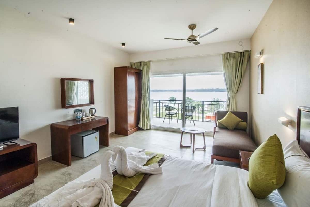 A hotel room with a bed, a desk, a television, a balcony with a view of the sea, and a ceiling fan.