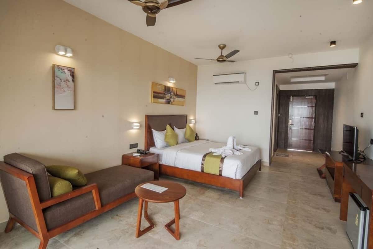 A hotel room with a bed, a sofa, a small table, a ceiling fan, and a television.