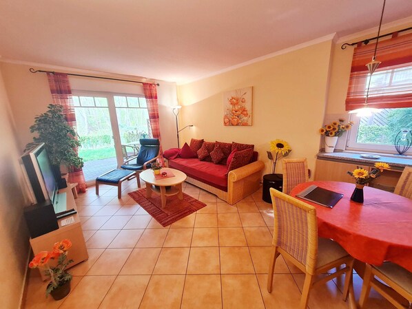 A cozy living room with a red sofa, a coffee table, and a dining table with chairs. There is a TV on a stand and a potted plant on the floor.