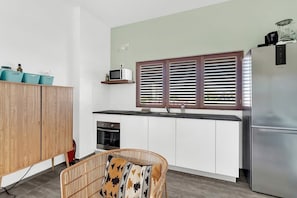 A compact kitchen with white cabinets, a stainless steel refrigerator, a microwave, and a window with blinds.