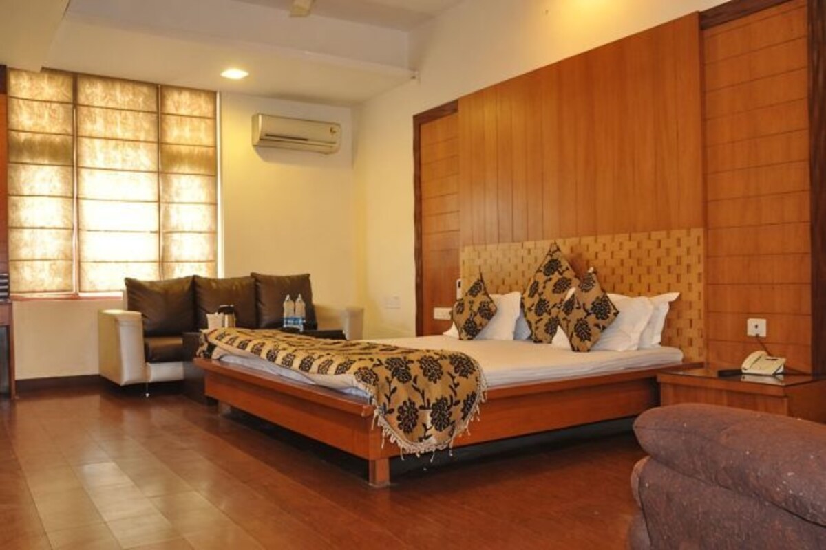 Una habitación de hotel con cabecera de madera, cama con ropa de cama estampada, mesita de noche, una silla y ventana con persianas.