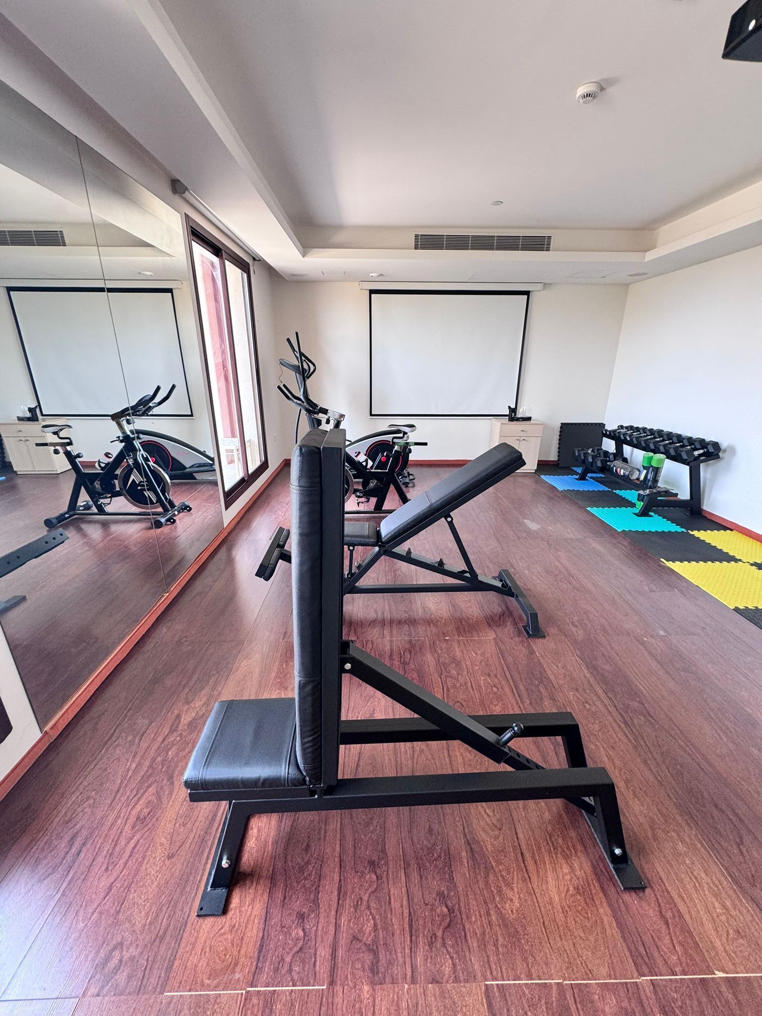 Un gimnasio con equipamiento de ejercicio, piso de madera y ventanas amplias.