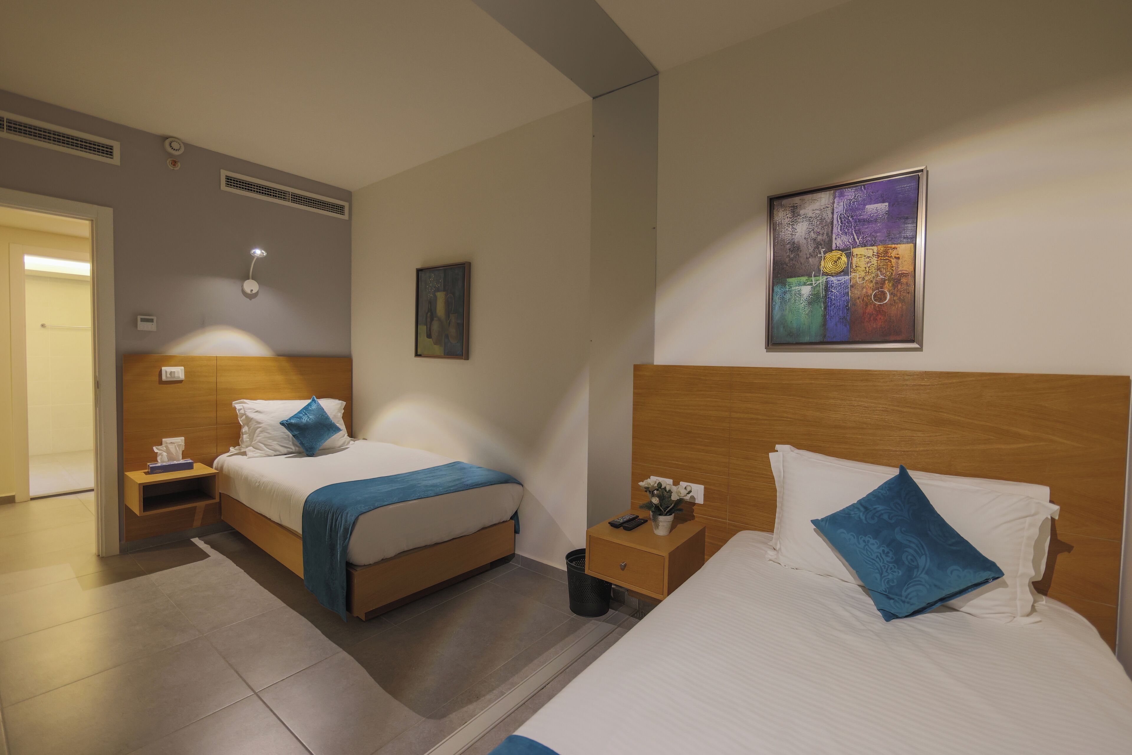 A hotel room with two beds, a nightstand, a wall-mounted lamp, and a painting.