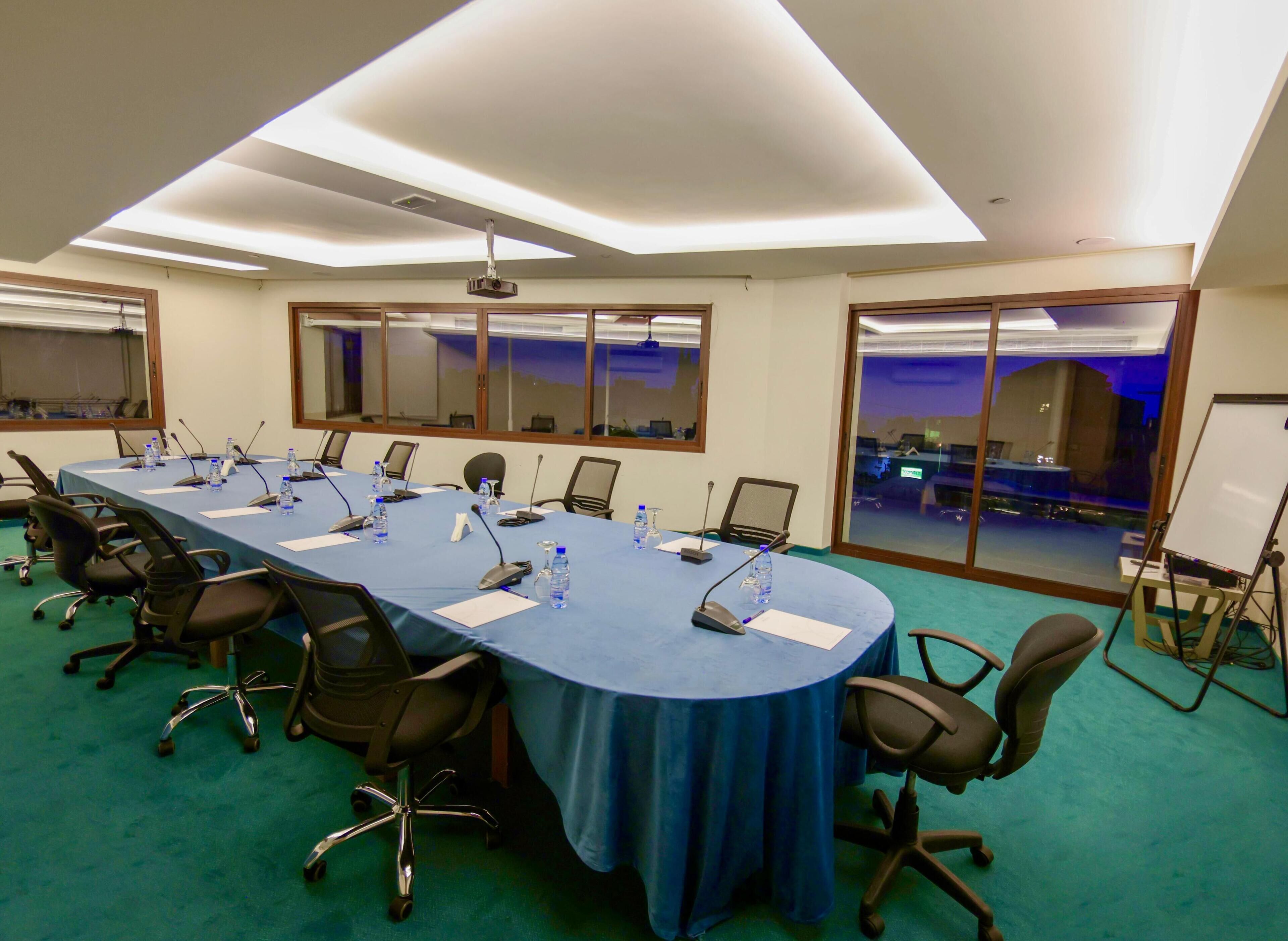 Una sala de conferencias con una mesa larga, sillas, micrófonos y un proyector.