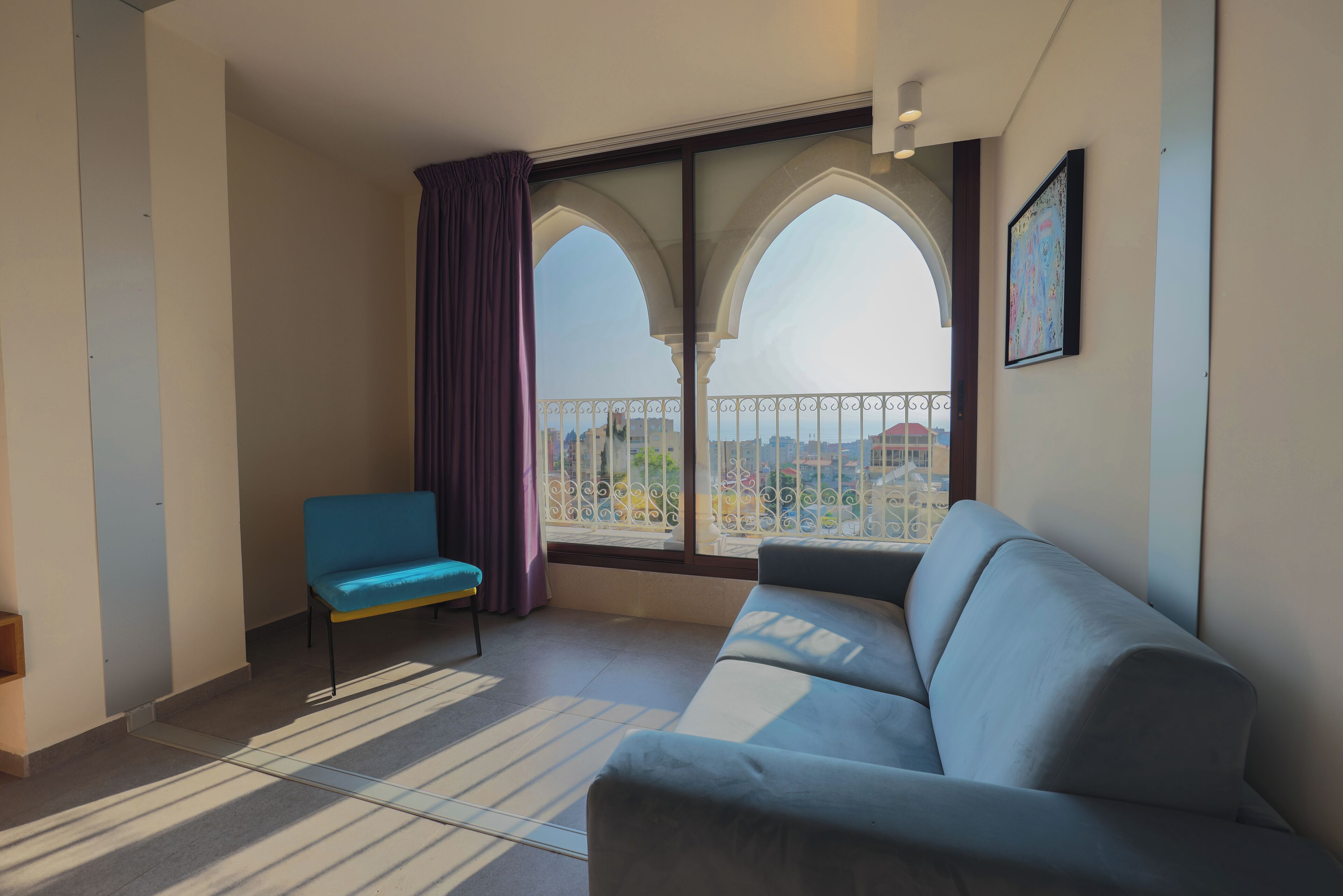 A modern living room with a sofa and a blue chair, a large window with a view of a cityscape, and a balcony with a railing.
