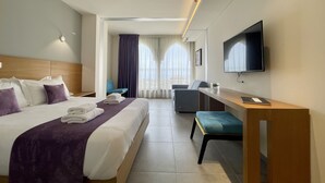 Een hotelkamer met een groot bed, een bureau, een stoel, een televisie en uitzicht op de stad door grote ramen.