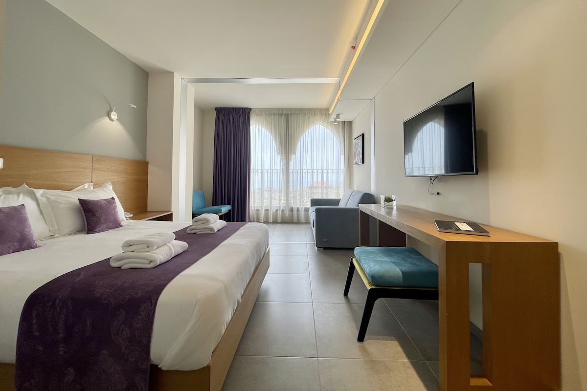A hotel room with a large bed, a desk, a chair, a TV, and a view of the city through large windows.
