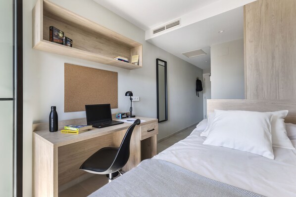 Una habitación de hotel moderna con un escritorio de madera, una computadora portátil, una silla negra, una cama con ropa blanca y un espejo en la pared.
