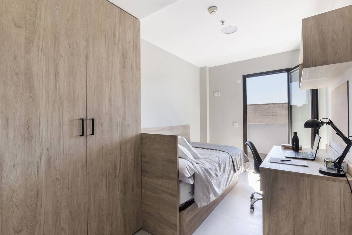 Een moderne slaapkamer met een houten garderobe, een bed met grijze beddengoed, een bureau met een laptop en een groot raam.