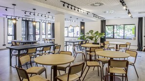 Een ruime eetruimte met meerdere tafels en stoelen, grote ramen en plafondlampen.