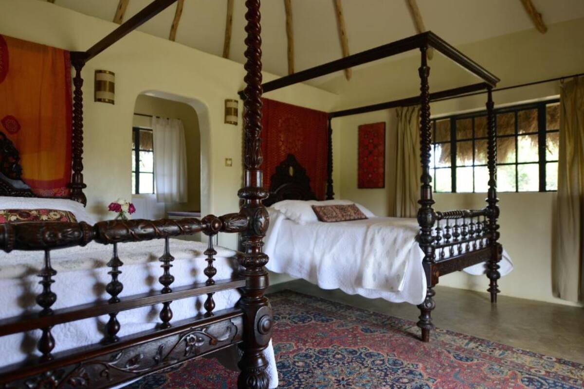 Un lit à baldaquin dans une pièce dotée d’un tapis à motifs, d’une grande fenêtre avec des rideaux et d’une tapisserie rouge au mur.