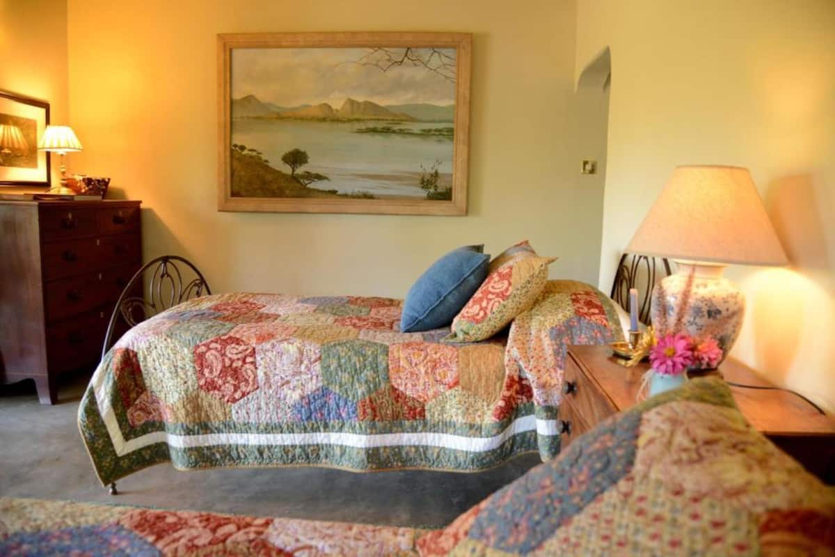 Une chambre à coucher avec un lit recouvert d’une couverture colorée, une commode en bois, une table de chevet avec une lampe et un tableau représentant un paysage accroché au mur.