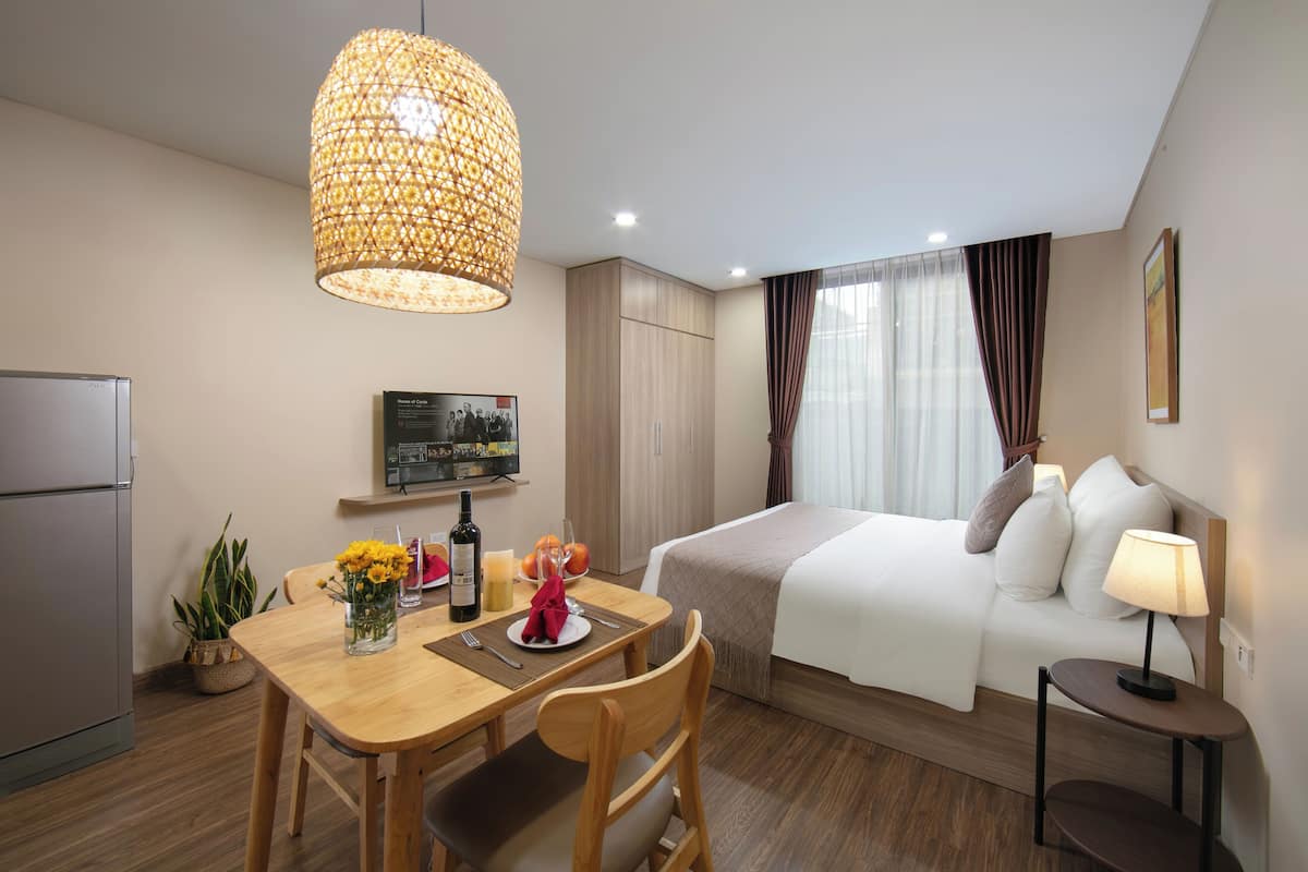 Habitación de hotel con cama, mesita de noche, lámpara, mesa de comedor con sillas, pequeña cocina y una lámpara colgante.