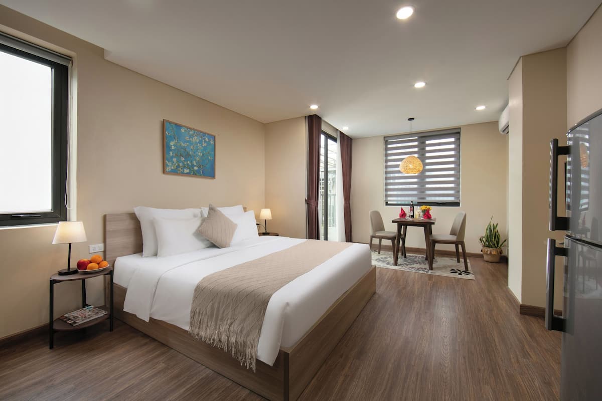 Habitación de hotel con una cama grande, una mesa de comedor con sillas, un refrigerador y una ventana con persianas.
