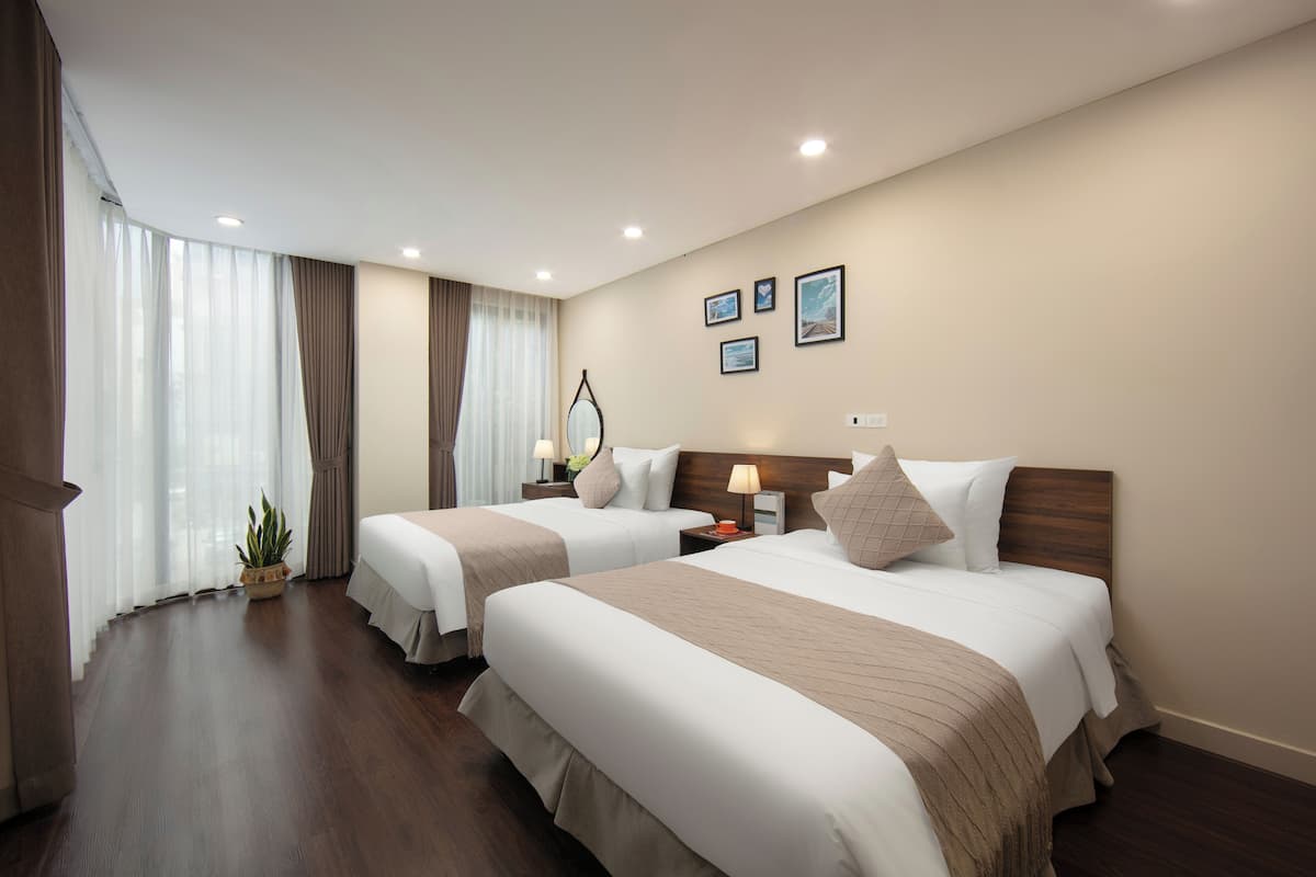 Habitación de hotel con dos camas, un ventanal con cortinas, una mesita de noche con lámpara y una planta pequeña.
