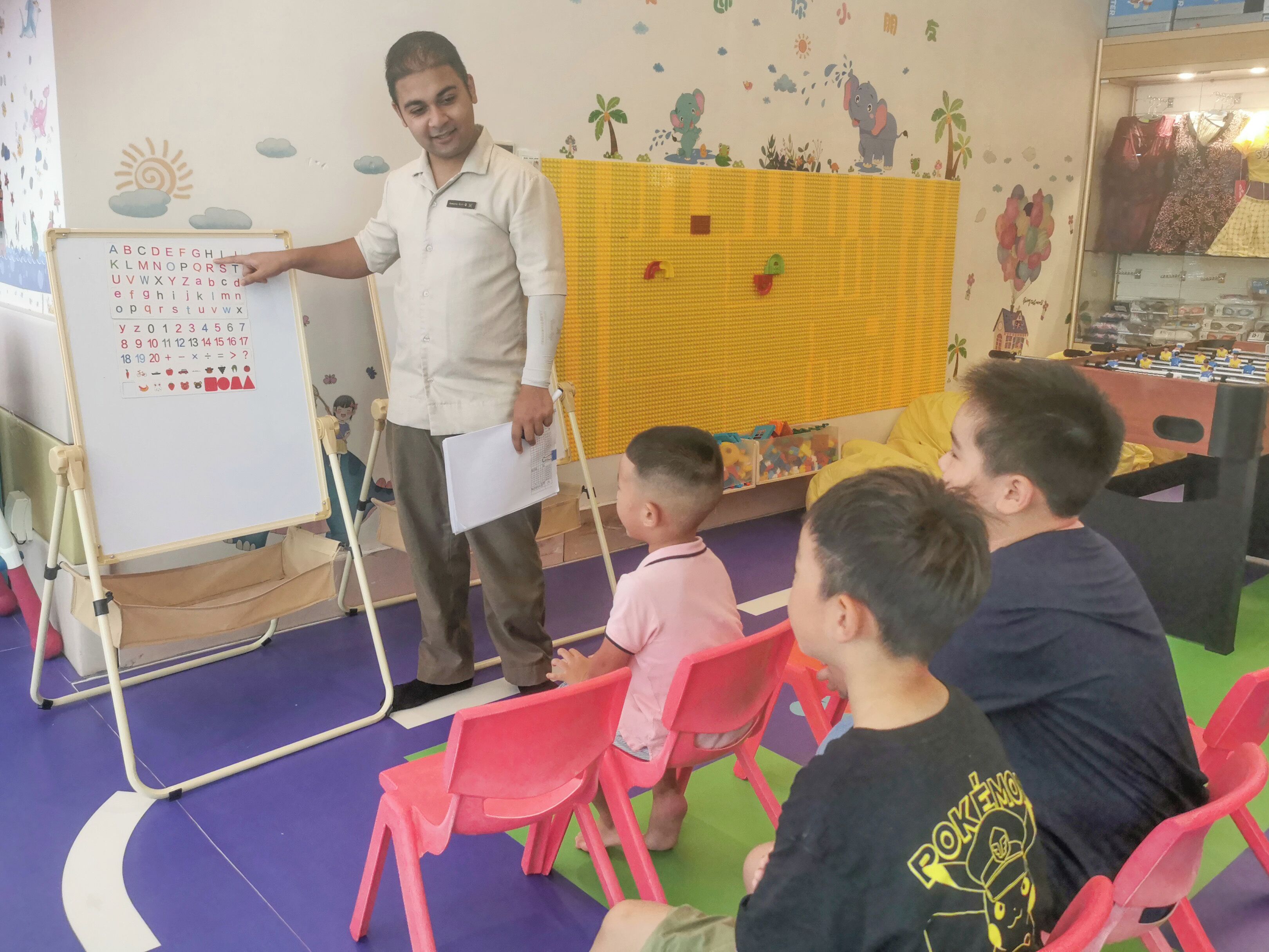 A classroom with a whiteboard displaying educational content, a teacher standing beside it, and children seated on chairs.
