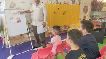A classroom with a whiteboard displaying educational content, a teacher standing beside it, and children seated on chairs.