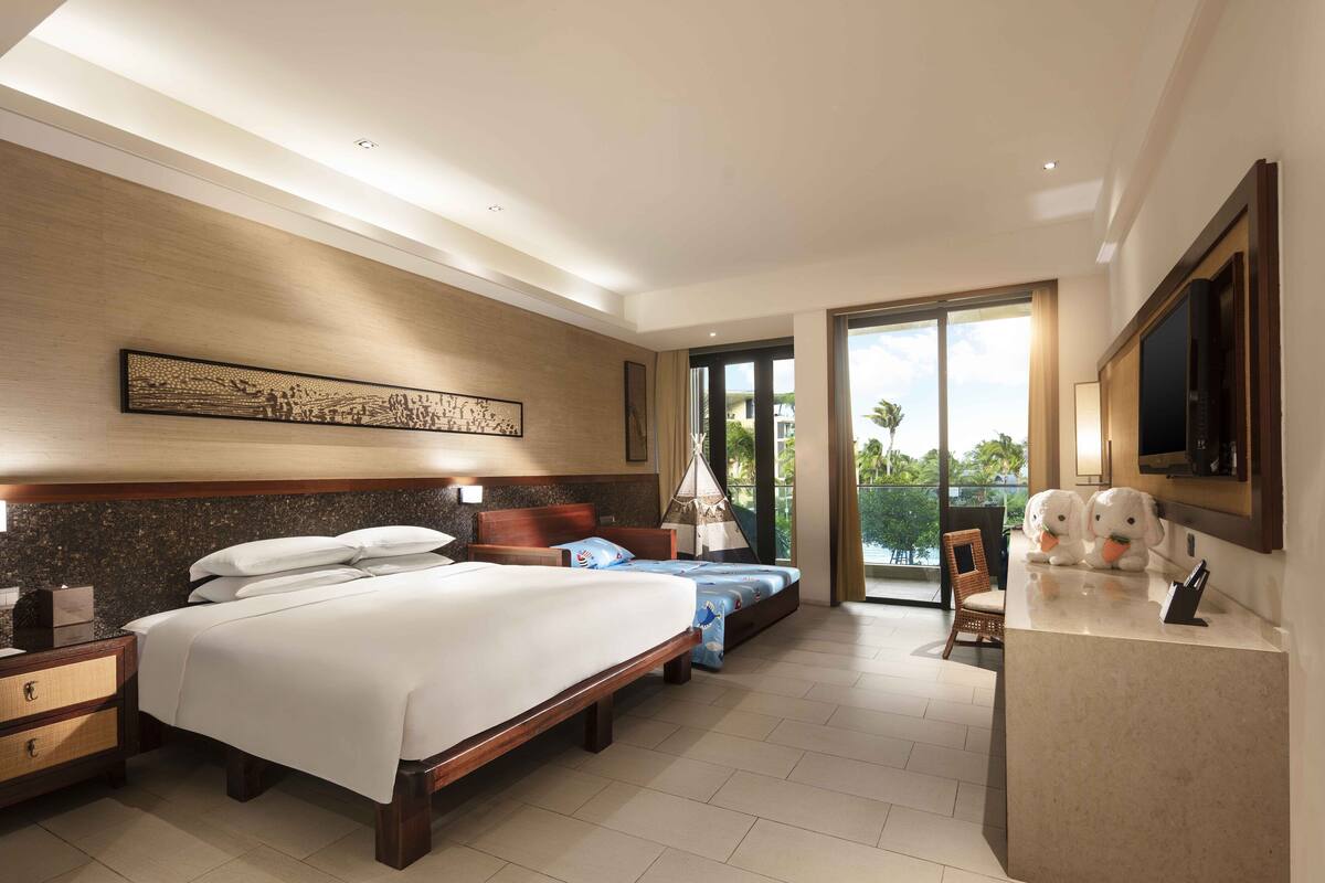 Une chambre d’hôtel avec un grand lit, un lit plus petit, un bureau avec une télévision et un balcon avec vue.