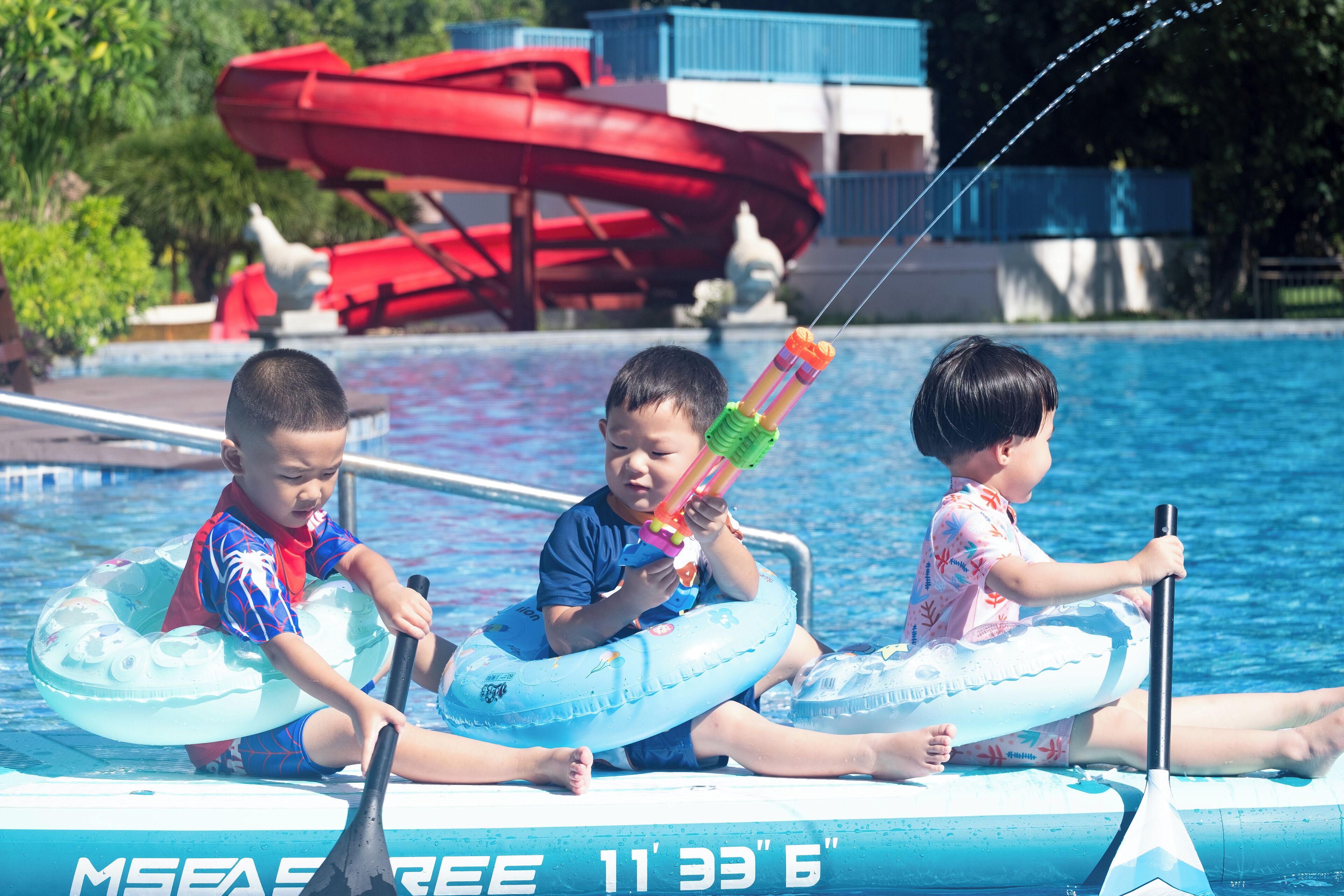 Trois enfants sur un flotteur de piscine, avec une pagaie, près d’un toboggan aquatique.