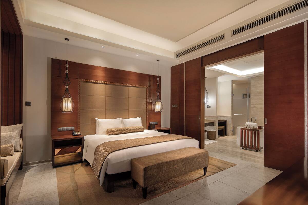 Une chambre d’hôtel moderne dotée d’un grand lit, d’un coin salon avec un banc, d’une table de chevet et d’une lampe fixée au mur.