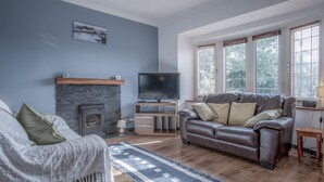 A living room with a fireplace, a leather sofa, a TV, and a large window with blinds. - Cambrian Cottage - 3 Bed Cottage - Tenby (Tenby)