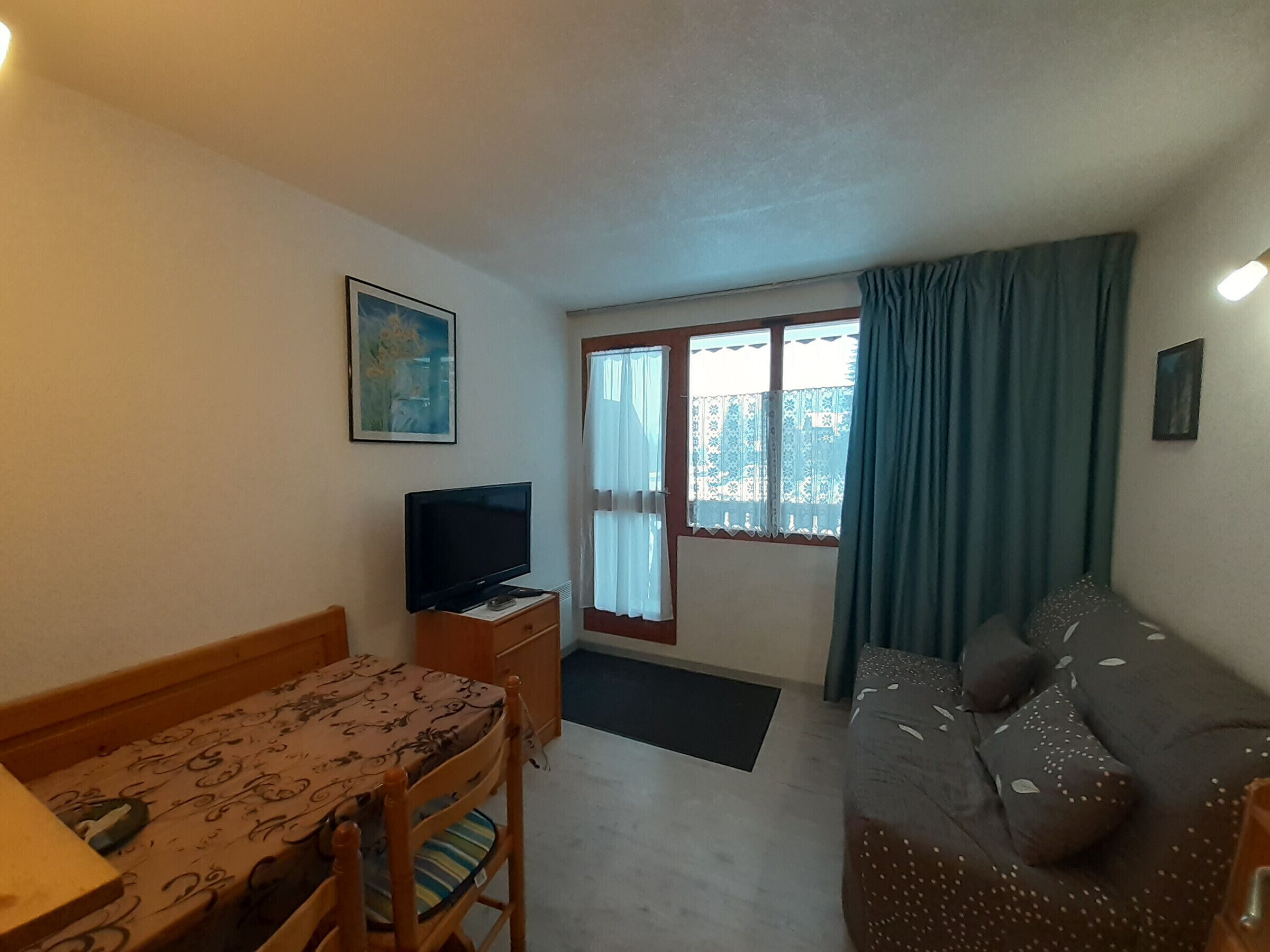 A small, well-lit room with a wooden table, a television, a sofa, and a window with curtains.