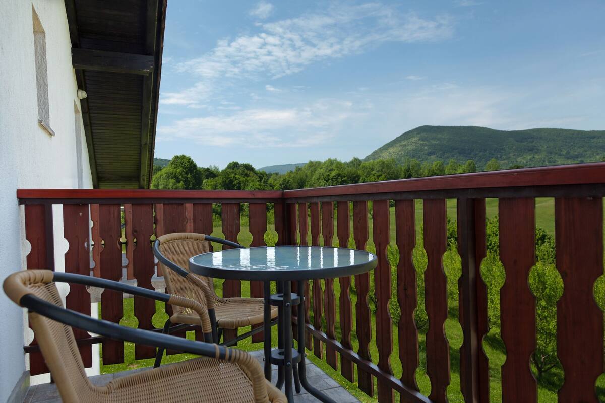 Ein Balkon mit Korbstühlen und einem Glastisch, mit Blick auf eine grüne Landschaft mit Hügeln.