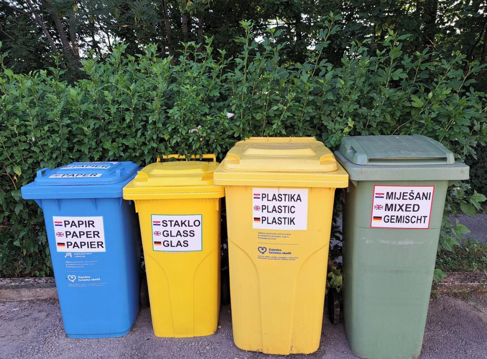 Quatre poubelles, étiquetées pour le papier, le verre, le plastique et les déchets mixtes, sont placées  à l’extérieur, avec une haie en arrière-plan.