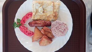 A plate with a pastry, sliced tomatoes, sausages, and a slice of meat on a white plate. - Broad Park Hotel (Mbale)
