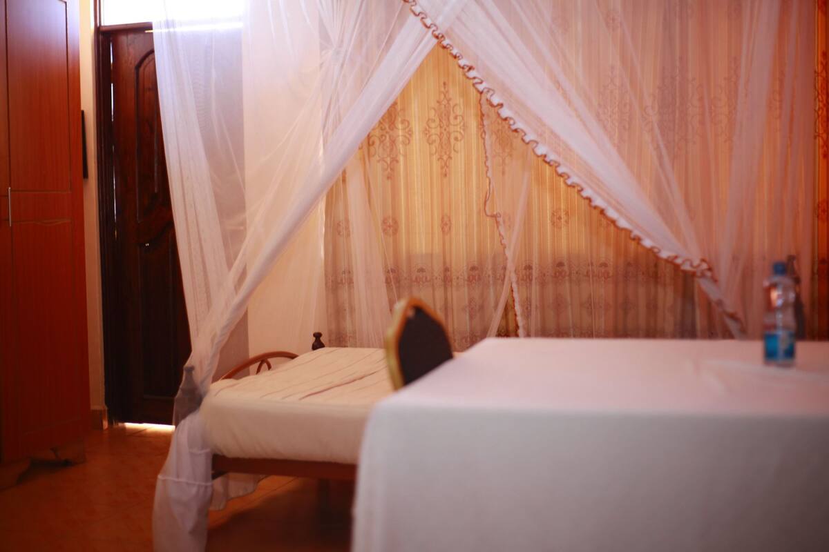 Quarto com cama sob um dossel, porta de madeira e uma mesa com uma garrafa.
