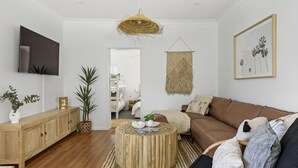 A modern living room with a wooden floor, a brown sofa, a round wooden coffee table, a TV mounted on the wall, and a hanging macrame wall hanging. - Berrara Beach House - Belle Escapes Jervis Bay (Berrara)