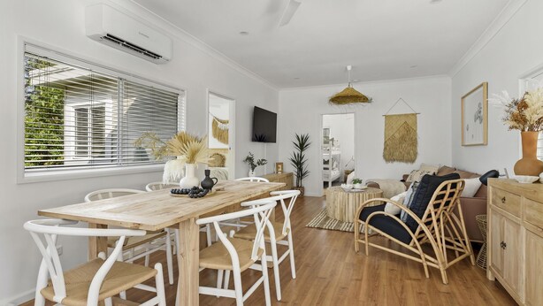 A spacious living room with a wooden dining table, white chairs, a sofa, and a TV mounted on the wall. - Berrara Beach House - Belle Escapes Jervis Bay (Berrara)