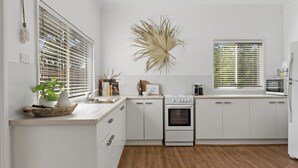 A modern kitchen with white cabinetry, a stove, and a sink. There are two windows with blinds, a decorative plant, and a framed picture on the counter. - Berrara Beach House - Belle Escapes Jervis Bay (Berrara)