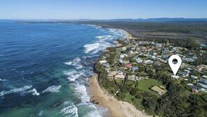 An aerial view of a coastal residential area with houses along the beach, a clear sky, and a marker indicating a specific location. - Berrara Beach House - Belle Escapes Jervis Bay (Berrara)
