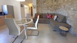 A modern living room with a stone wall, a grey sofa, a glass coffee table, and a dining area with chairs. - Holiday house on peaceful location in the suburb of Pula (Pula)
