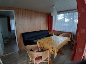 Una habitación con pared de paneles de madera, un sofá negro, un sillón de mimbre, una mesa con mantel floral y una ventana con cortinas rojas.
