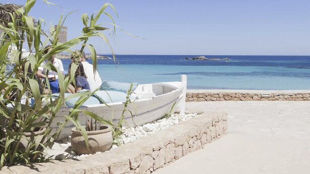 A beachside view with a boat, a stone wall, and a clear blue sea. - Roquetes Rooms - Formentera Break (Formentera)