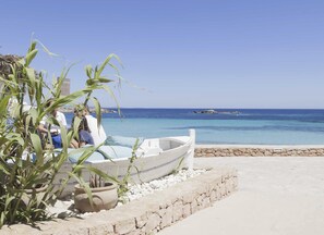 A beachside view with a boat, a stone wall, and a clear blue sea. - Roquetes Rooms - Formentera Break (Formentera)